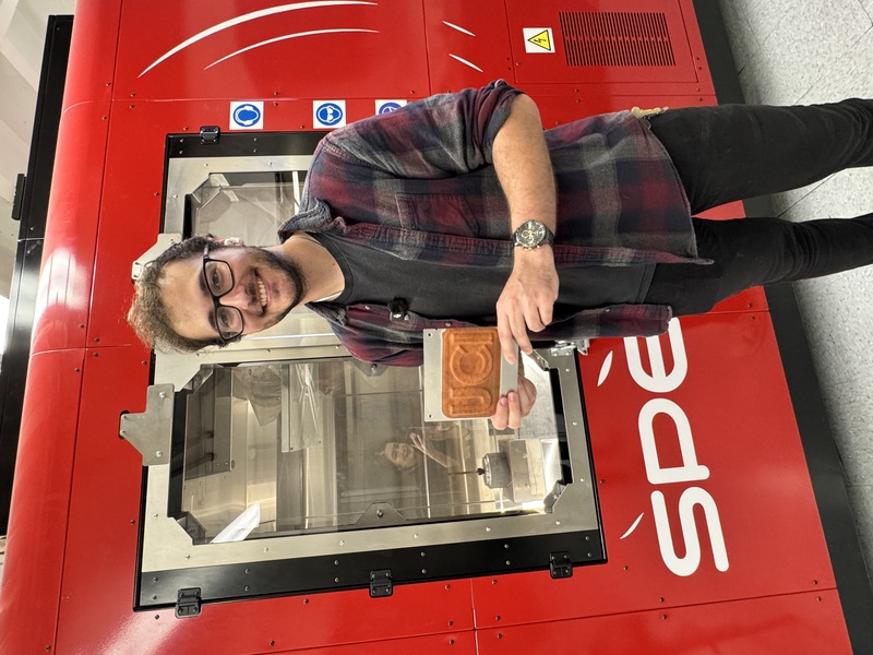 UCI doctoral student Michael Ross holds up a UCI metal plate made by the million-dollar cold spray machine behind him. (Photo: Natalie Tso/UCI)